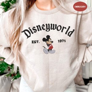 A person wearing a beige sweater with the text "Disneyworld EST. 1971" and a Mickey Mouse graphic printed on it.