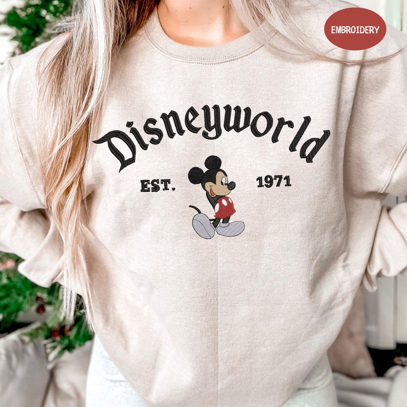 A person wearing a beige sweater with the text "Disneyworld EST. 1971" and a Mickey Mouse graphic printed on it.