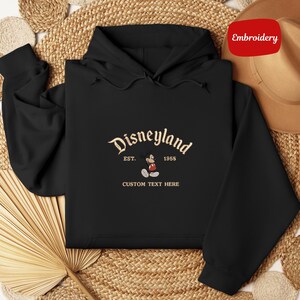 A black hoodie with a Disneyland logo and text, along with a hat and a woven mat, are displayed in the image.