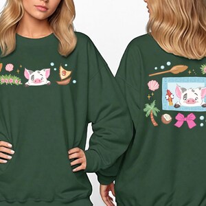 two women wearing green sweatshirts with colorful and whimsical designs, including a pig, flowers, and other decorative elements.