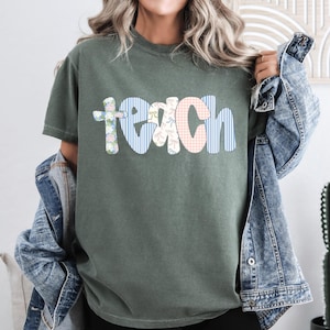May include: An olive green t-shirt featuring the word "TEACH" in patchwork lettering. The letters are adorned with floral, striped, and gingham patterns. A denim jacket is worn over the shoulders.