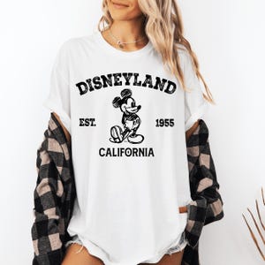 A woman wearing a white t-shirt with a graphic of Mickey Mouse and text that says "Disneyland EST. 1955 CALIFORNIA".