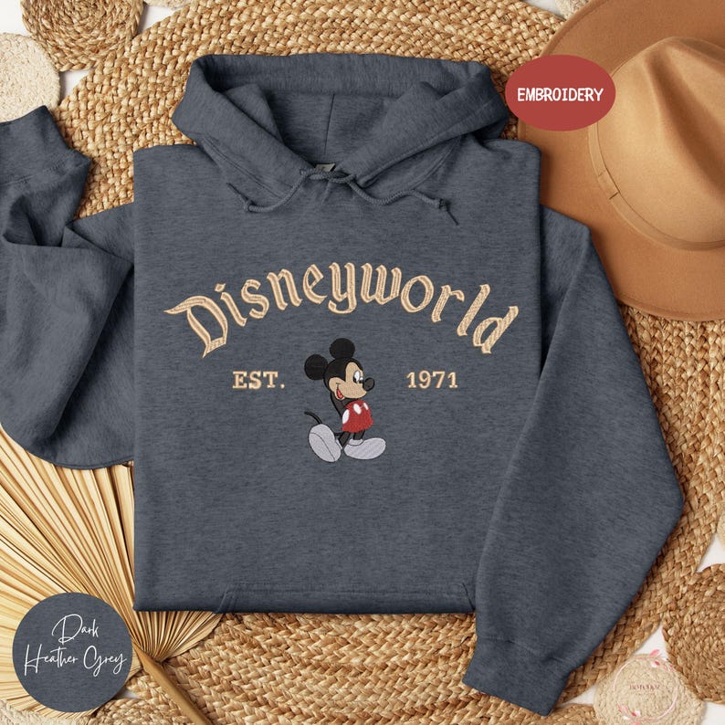 A gray hoodie with the text "Disneyworld EST. 1971" and a Mickey Mouse graphic, along with a straw hat and woven mat, are displayed in the image.