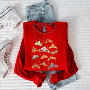 A red sweater with a design of colorful crowns and tiaras, a pair of denim jeans, and white sneakers are laid out on a white surface.