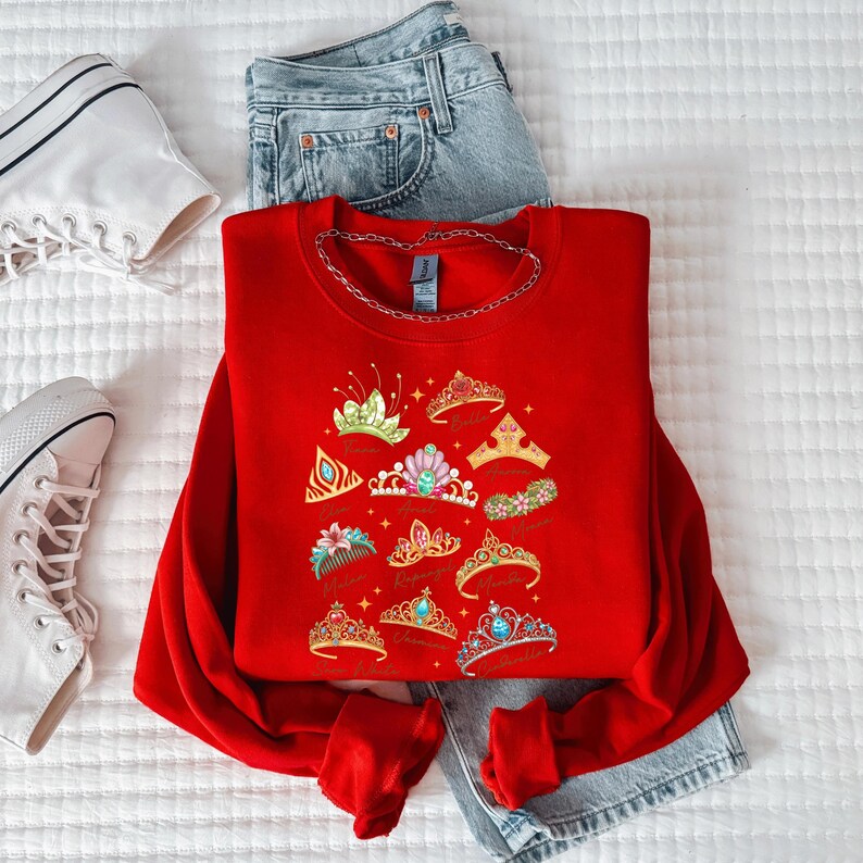 A red sweater with a design of colorful crowns and tiaras, a pair of denim jeans, and white sneakers are laid out on a white surface.
