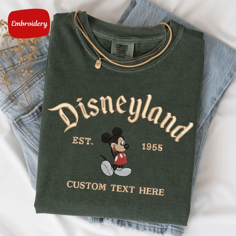 A green t-shirt with a Mickey Mouse embroidered on it, along with a tag that says "Disneyland EST. 1955" and a tag that says "COMFORT COLORS". The t-shirt is placed on a white surface, and there are some jeans visible in the background.