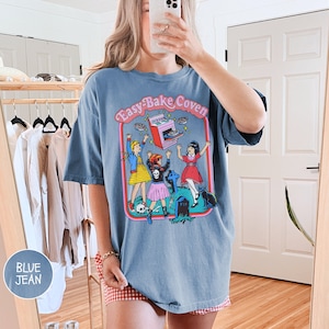 Pu&ograve; includere: Una maglietta oversize in jeans blu con una stampa grafica di tre ragazze davanti a una lapide con il testo "Easy-Bake Coven" e un forno Easy-Bake volante.