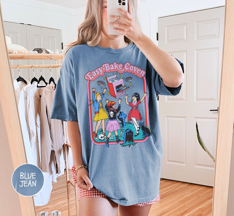 Pu&ograve; includere: Una maglietta oversize in jeans blu con una stampa grafica di tre ragazze davanti a una lapide con il testo "Easy-Bake Coven" e un forno Easy-Bake volante.