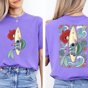 a woman wearing a purple t-shirt with a graphic design featuring a mermaid and a surfboard.