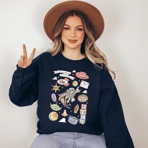 A woman wearing a black sweatshirt with a graphic design of various cartoon characters and icons, including a character holding a sign and a character holding a sign with a heart.