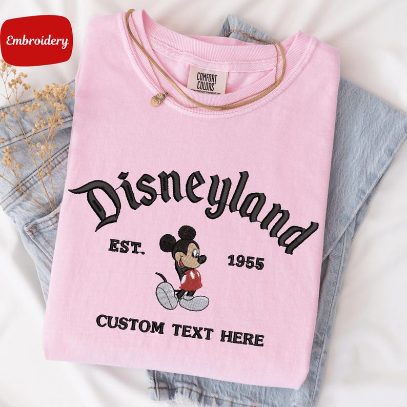 A pink t-shirt with a Mickey Mouse design and text, along with a necklace, is displayed on a white surface.
