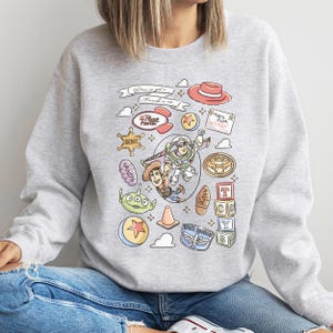 A person wearing a gray sweatshirt with a colorful graphic design featuring various characters and objects from the Toy Story franchise.