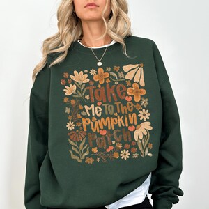 May include: Dark green sweatshirt featuring the text "Take Me To The Pumpkin Patch" in orange and brown, framed by floral designs in orange, cream, and brown. The sweatshirt has a white collar.