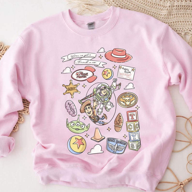A pink sweatshirt with a cartoon-style illustration of various characters and objects from the Toy Story franchise, including Woody, Buzz Lightyear, and other iconic figures from the movie.