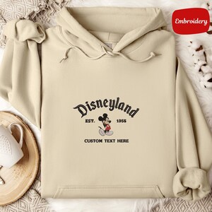 A beige hoodie with a Disneyland logo and text, along with a mug and a white cloth, arranged on a surface.