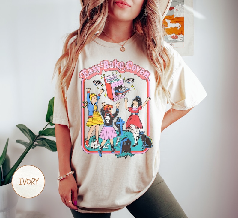 Pu&ograve; includere: Una maglietta color crema con una stampa grafica di tre ragazze davanti a una lapide e un forno Easy-Bake. Il testo "Easy-Bake Coven" &egrave; stampato sopra la grafica.