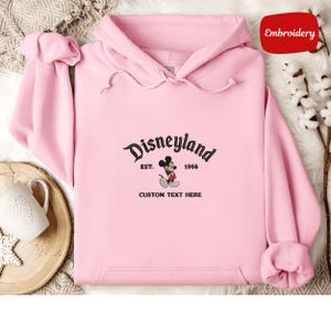 A pink hoodie with a Disneyland logo and text, along with a mug and some cotton balls, is displayed on a surface.
