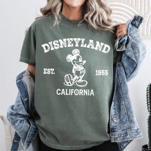 A woman wearing a green t-shirt with a Mickey Mouse graphic and text, standing in front of a denim jacket.