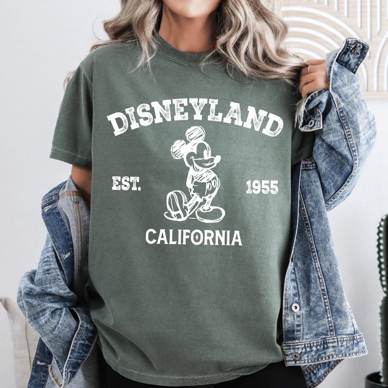 A woman wearing a green t-shirt with a Mickey Mouse graphic and text, standing in front of a denim jacket.