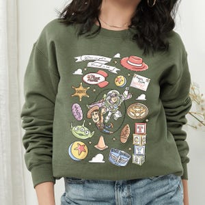 A green sweatshirt with a collage of various cartoon characters and icons, including Woody from Toy Story, Buzz Lightyear, and other iconic figures from the Toy Story franchise.