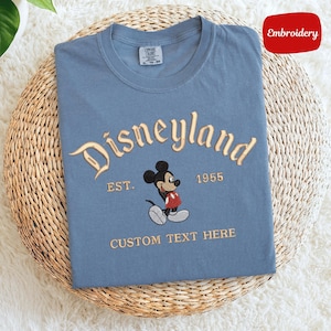 A blue t-shirt with a Mickey Mouse design and text that says "Disneyland EST. 1955 CUSTOM TEXT HERE" is displayed on a woven basket.