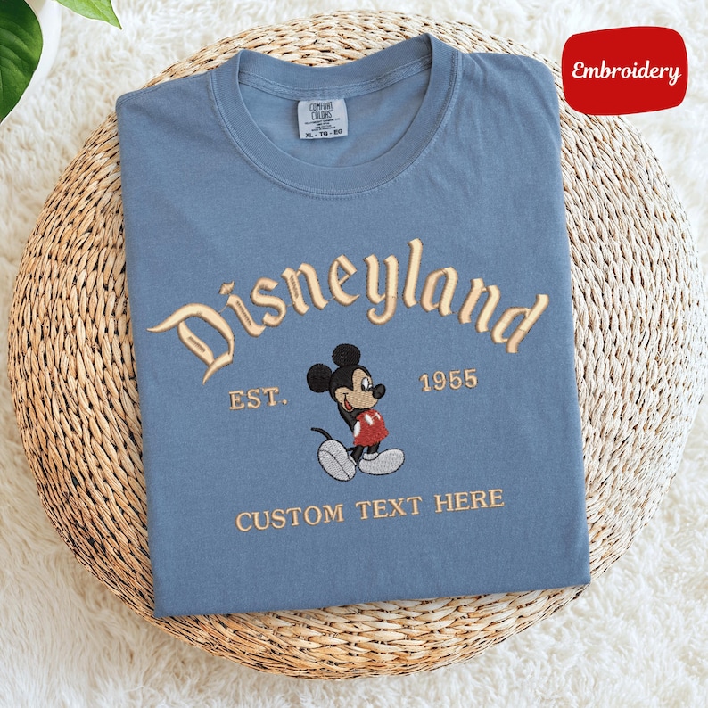 A blue t-shirt with a Mickey Mouse design and text that says "Disneyland EST. 1955 CUSTOM TEXT HERE" is displayed on a woven basket.