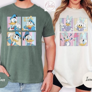 May include: Two t-shirts, one moss green and one ivory, each featuring a cartoon character design. The moss green shirt has Donald Duck in four different poses, while the ivory shirt has Daisy Duck in four different poses. Both shirts are short-sleeved.