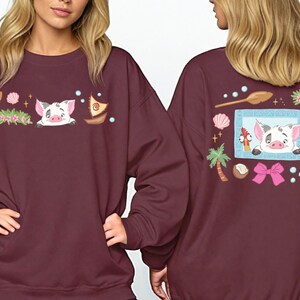 two women wearing burgundy sweatshirts with colorful and whimsical designs, including a pig, flowers, and other decorative elements.