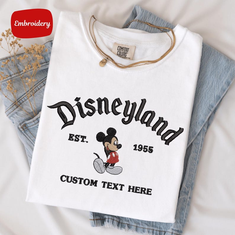 A white t-shirt with a Mickey Mouse design and text, along with a denim skirt, is displayed on a white surface.