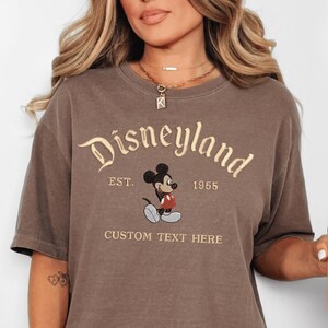 A woman wearing a brown t-shirt with a Mickey Mouse design and text that says "Disneyland EST. 1955 CUSTOM TEXT HERE".
