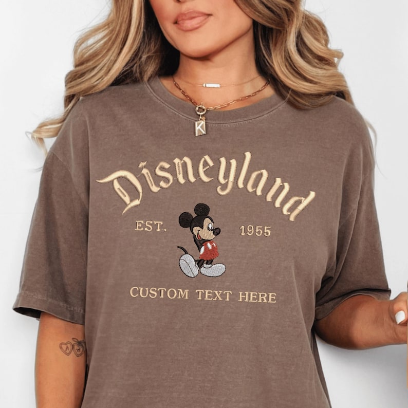 A woman wearing a brown t-shirt with a Mickey Mouse design and text that says "Disneyland EST. 1955 CUSTOM TEXT HERE".