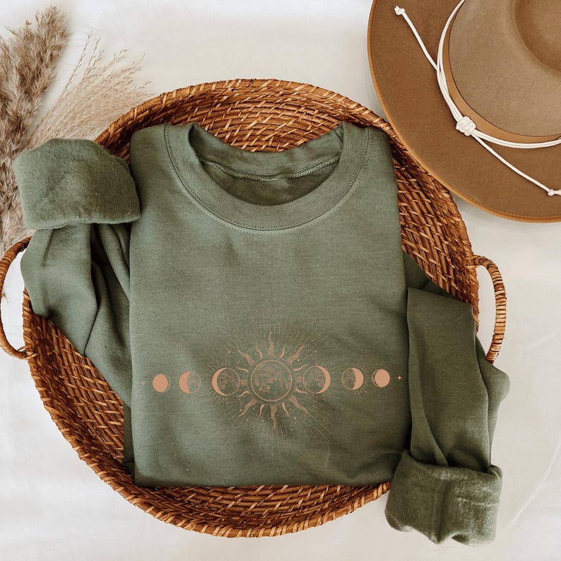 a green sweatshirt with a sun and moon design, a brown hat, and a woven basket containing a green sweater and a pair of pants.