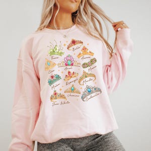 A woman wearing a pink sweatshirt with a graphic design of various princess crowns and titles.