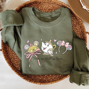 May include: An olive green sweatshirt with a graphic of a teapot, a fairytale character, and mouse ears with a pink bow. The text "Tale as old as time" is printed below. The sweatshirt is folded in a woven basket.