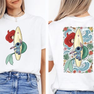 A woman wearing a white t-shirt with a colorful graphic of a mermaid and a surfboard on the back.