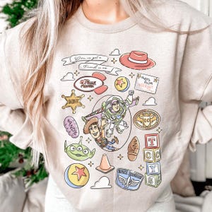 a person wearing a light-colored sweater with a collage of various cartoon characters and icons, including Woody from Toy Story, Buzz Lightyear, and other iconic characters from the Toy Story franchise.