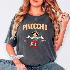 May include: A dark gray t-shirt with a vintage-style graphic of Pinocchio, the Disney character. The graphic features Pinocchio in his signature red shorts and blue shirt, with the text "PINOCCHIO EST. 1940" above and below him.