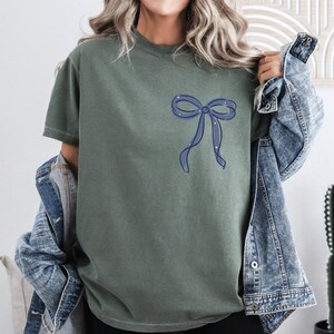 A woman wearing a green t-shirt with a blue bow on it, paired with a denim jacket, stands in front of a white wall.