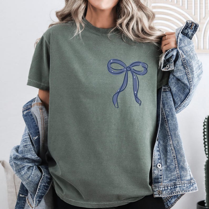 A woman wearing a green t-shirt with a blue bow on it, paired with a denim jacket, stands in front of a white wall.