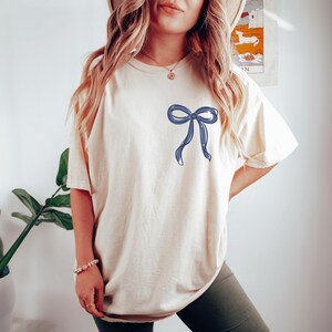A woman wearing a white t-shirt with a blue bow on it, standing in front of a plant and a poster on the wall.
