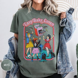 Pu&ograve; includere: Una maglietta verde con una stampa grafica di tre ragazze davanti a un forno con il testo "Easy-Bake Coven".