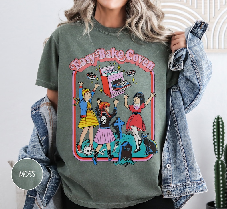 Pu&ograve; includere: Una maglietta verde con una stampa grafica di tre ragazze davanti a un forno con il testo "Easy-Bake Coven".
