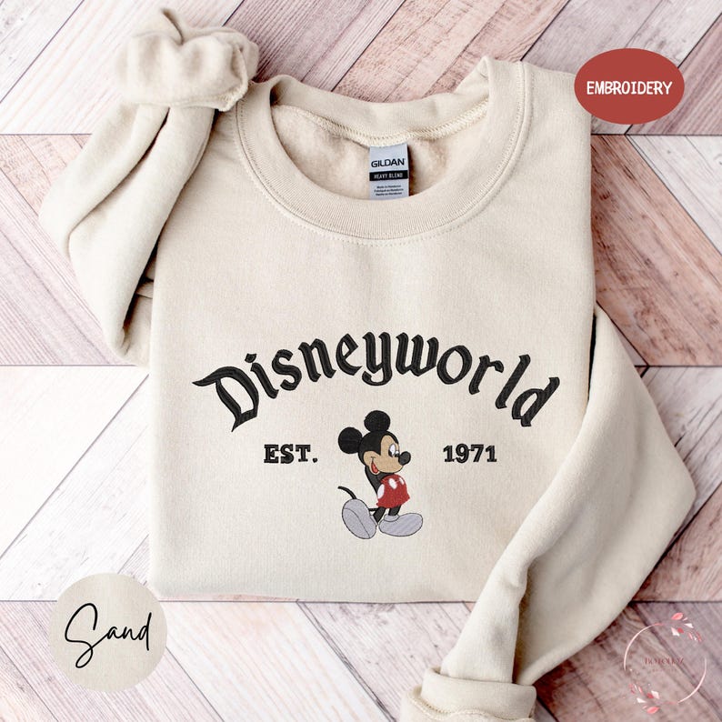A beige sweater with the text "Disneyworld EST. 1971" and a Mickey Mouse graphic printed on it, laid out on a wooden surface.