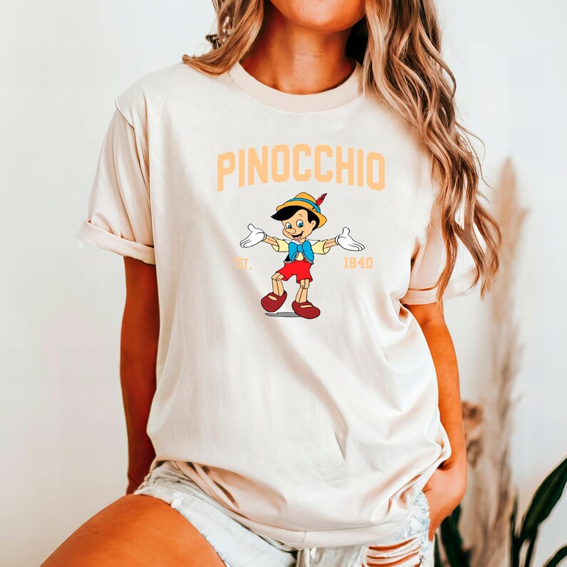May include: A beige t-shirt with a graphic of Pinocchio from the 1940 Disney film. The graphic is in orange and features Pinocchio with his arms outstretched. The text "PINOCCHIO EST. 1940" is printed in orange above the graphic.