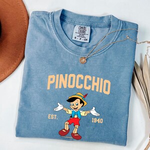 May include: A light blue t-shirt with a vintage-style graphic of Pinocchio, a cartoon character from the Disney movie. The graphic includes the text "Pinocchio", "Est. 1940", and a cartoon image of Pinocchio.
