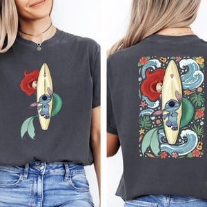 a woman wearing a gray t-shirt with a colorful graphic design on the back. The design depicts a mermaid riding a surfboard, surrounded by various elements such as flowers, waves, and a starfish.