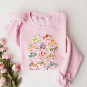 A pink sweatshirt with a graphic design of various princess crowns and tiaras printed on it, along with the names of the princesses.