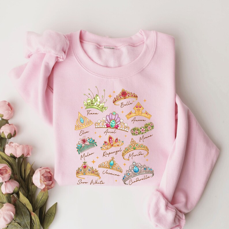 A pink sweatshirt with a graphic design of various princess crowns and tiaras printed on it, along with the names of the princesses.
