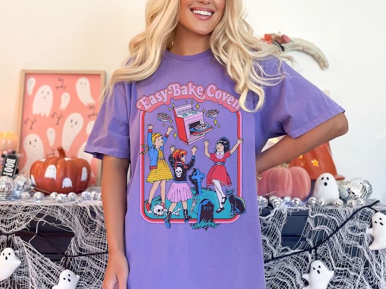 Pu&ograve; includere: Maglietta viola con una grafica di tre ragazze davanti a un forno Easy-Bake rosa. Le ragazze indossano costumi di Halloween e il forno &egrave; in levitazione. Il testo "Easy-Bake Coven" &egrave; sopra il forno.