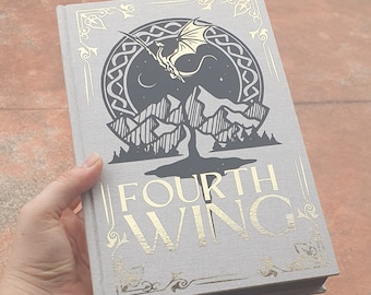 Op de afbeelding: Een grijs hardback boek met een gouden ontwerp op de omslag. Het ontwerp toont een zwart silhouet van een draak die over een bergketen vliegt met een halve maan en sterren op de achtergrond. De titel "FOURTH WING" is in gouden letters op de voorkant gedrukt.